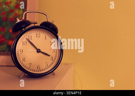 Classic black retro alarm clock with twin bells standing on a white surface, set against a warm yellow background, symbolizing morning, deadlines, and Stock Photo