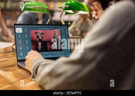 Student uses a laptop AI image generator interface during collaborative study at a library table, with classmates, books and green lamps around the desk. Stock Photo