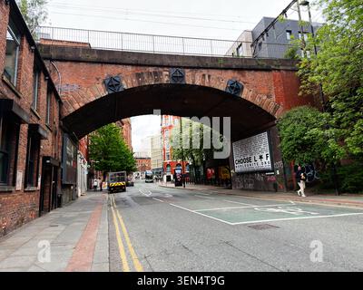 Manchester, UK - May 07, 2026: Historic red brick arch spans an urban street in Manchester, UK with buses, pedestrians and graffiti. Stock Photo