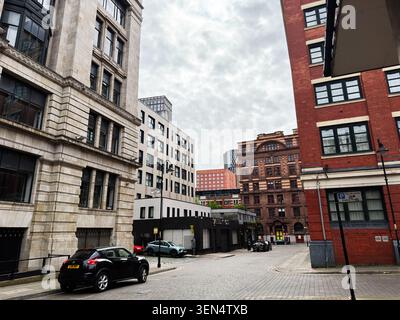 Manchester, UK - May 07, 2026: Cobblestone street in Manchester UK lined with historic and modern buildings, parked cars and cloudy sky. Stock Photo