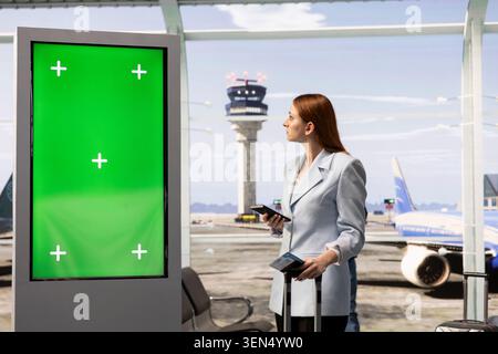Green screen display with woman traveler with luggage looking at huge board in terminal, checking boarding pass in hand and waiting for departure. Chroma key screen for advertising. Stock Photo