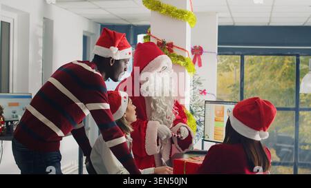Manager dressed as Santa Claus in xmas ornate office using candy cane ornament as pointer while showing numbers on screen. Supervisor wearing Christmas costume overseeing work done by his team Stock Photo
