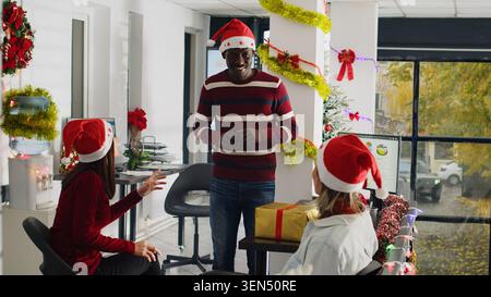 Company personnel wearing Santa hat enjoying Christmas spirit mood, playing pretending game during shift break at work. Colleagues having fun together during winter holiday season in xmas adorn office Stock Photo