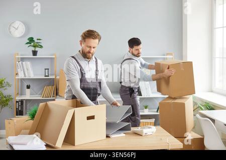 Moving office service, two men in overalls packing boxes, carrying items, placing belongings on desk Stock Photo