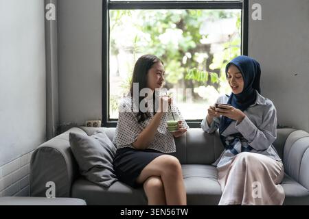 Two women chatting on a sofa while sharing a drink and looking at a smartphone near a bright window Stock Photo