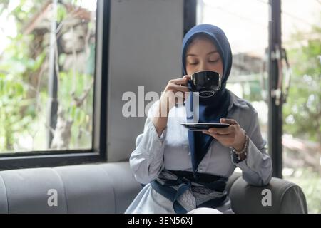 Woman in hijab drinking coffee and using smartphone while sitting on a sofa near a window Stock Photo
