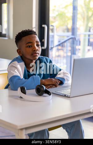 African American child boy wearing blue sweater and pants sitting at desk using laptop, headphones Stock Photo