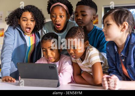 Diverse school-age children gathering around tablet in folio case at school table, watching screen Stock Photo