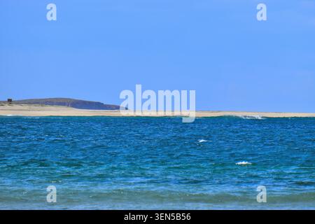 Clear blue water rolls in gentle waves toward a pale sandy dune, with a rocky landform rising in the distance beneath a bright sky. Stock Photo