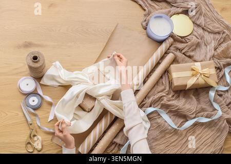 Overhead view of hands wrapping gift box with brown paper and ribbons on wooden table for special occasions, crafting supplies like scissors, thread Stock Photo