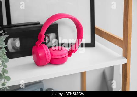 Bright pink wireless headphones on white shelf with retro VHS tapes and modern decor, music and technology, this arrangement combines elements of past Stock Photo