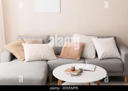 Modern gray sectional sofa with cozy pillows and coffee table in a minimalist living room interior, a white coffee table in front of the sofa holds bo Stock Photo