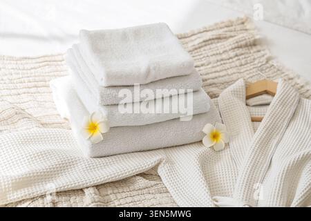 Stacked white terry cloth towels and waffle weave robe with plumeria flowers for spa and relaxation, this arrangement suggests a serene spa or relaxat Stock Photo