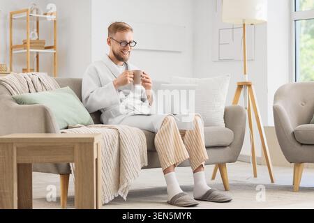 Smiling man in robe and pajamas working remotely on laptop with coffee at home, comfortable lifestyle and freelance work Stock Photo