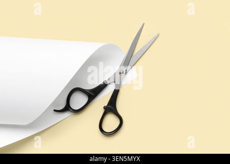Black handled scissors with white paper roll on light yellow background, crafting and office supplies Stock Photo