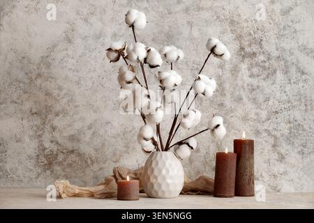 Natural cotton stems in white vase with lit brown candles and fabric on textured background, three lit brown pillar candles and a draped piece of ligh Stock Photo