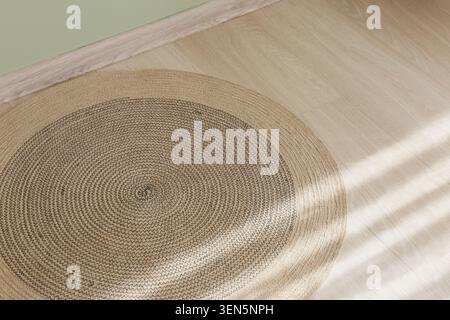 Round woven jute rug on light wood floor with sunlight and green wall baseboard, home decor and interior design Stock Photo