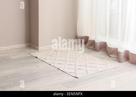 Neutral beige diamond patterned area rug on light wood floor with sheer white curtains and wall corner, sheer white curtains with a subtle brown trim Stock Photo