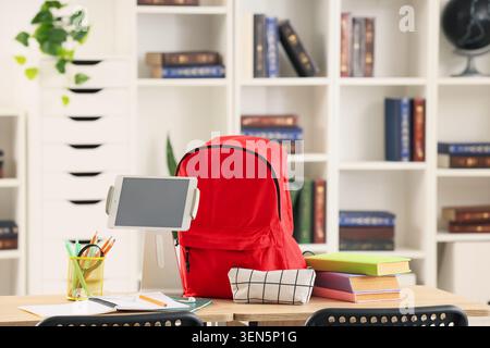 Red backpack, digital tablet, and school supplies on wooden desk in modern classroom or home study area Stock Photo