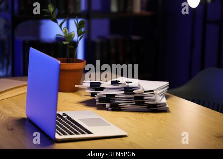 Late night office work with laptop, stacked documents, and potted plant on wooden desk in dark room Stock Photo