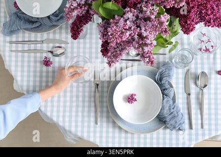 Elegant dining table setting with lilac flowers and checkered tablecloth, overhead view of hand placing glass Stock Photo