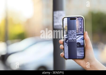 Hand holding smartphone scanning QR code on city street pole, digital information access and modern technology, the smartphone screen displays the QR Stock Photo