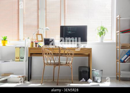 Modern home office workspace with computer monitor displaying code and laptop on wooden desk, a rattan chair sits in front of the desk, and a window w Stock Photo