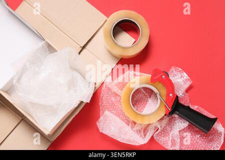 Cardboard box with bubble wrap, packing tape, and dispenser on red background for shipping and moving Stock Photo