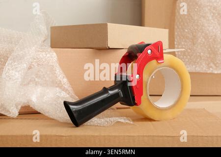 Packing tape dispenser with clear adhesive tape roll on cardboard boxes and bubble wrap for moving and shipping, the scene suggests the process of sec Stock Photo