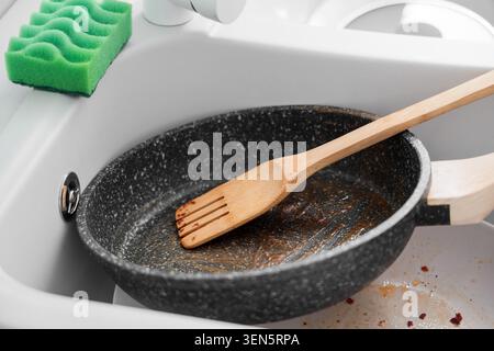 Dirty frying pan with wooden spatula and green sponge in kitchen sink after cooking meal, a green cleaning sponge sits on the edge of the sink, ready Stock Photo