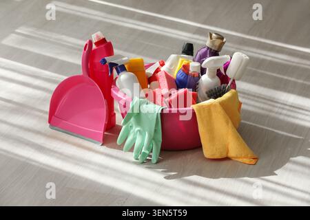 Household cleaning supplies and equipment in pink bucket with dustpan, gloves, and towel on light wood floor Stock Photo