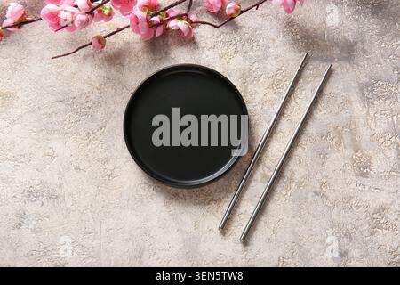 Empty black plate with metal chopsticks and pink cherry blossom branch on textured concrete background Stock Photo