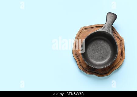 Empty black cast iron skillet on wooden cutting board, kitchen utensil for cooking and serving food, isolated on blue background Stock Photo