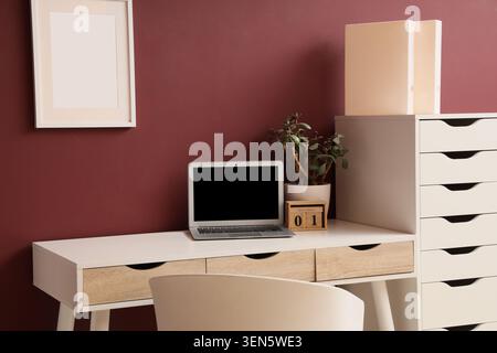 Modern home office workspace with laptop, desk, drawers, plant, and calendar against a burgundy wall Stock Photo