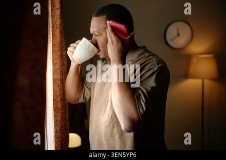 Black Adult Man Drinking Coffee While Using Smartphone at Home Stock Photo