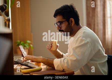 Adult Indian Man Eating While Using Laptop at Home for Work Stock Photo