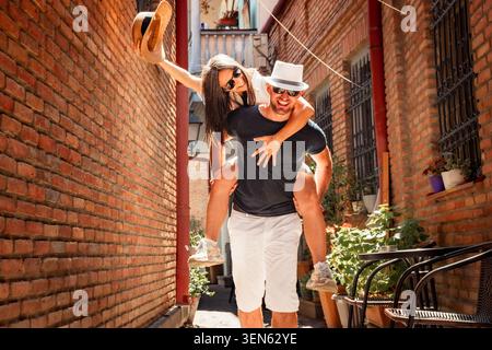 Happy interracial couple having fun piggyback ride in Tbilisi old city narrow street.Cheerful young man giving girlfriend piggyback ride.Couple laughi Stock Photo