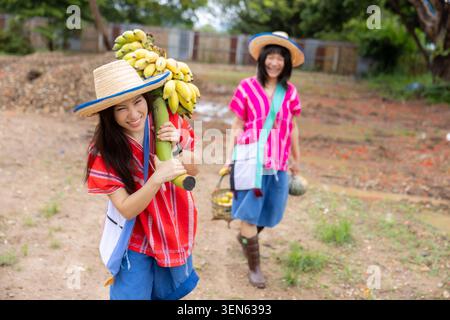 Banana woman basket hat rural farm smile tropical fruit village two smiling woman carrying banana bunch and basket outdoors on dry ground with tree an Stock Photo