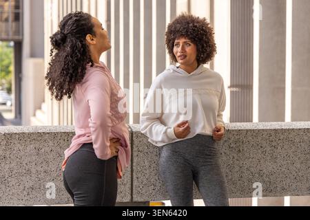 Diverse female friends chatting and resting on balcony by concrete railing, in hoodies and leggings Stock Photo