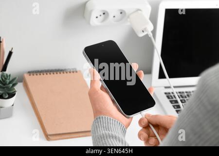 Person connecting smartphone to charger on desk with laptop and power strip, modern technology and device charging Stock Photo
