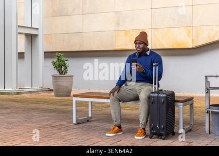 African American man in his 20s sitting on bench at plaza holding phone, suitcase, copy space Stock Photo