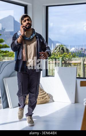 Bearded man in beanie scarf, holding phone while walking in studio with canvases and marina view Stock Photo