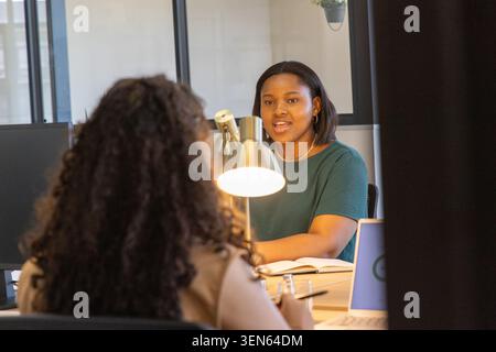 Diverse female colleagues discussing project at office desk with metal lamp, laptop, open notebook Stock Photo