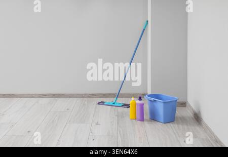 Floor cleaning supplies with mop, bucket, and detergents in empty room with gray walls and light wood floor Stock Photo
