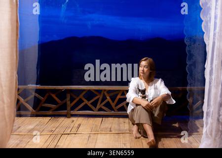 Thoughtful woman holding cute cat sits on wooden balcony terrace overlooking deep blue night mountain landscape framed by white curtain, expressing se Stock Photo