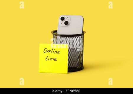 Digital detox with smartphone in trash can and "offline time" note on yellow background, a yellow sticky note with "offline time" handwritten on it re Stock Photo