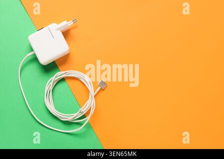 White laptop charger and coiled cable on green and orange background, power adapter for electronic devices, the power adapter is ready for use with el Stock Photo