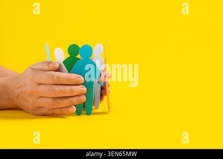Human hands protecting diverse paper people chain on yellow background community support and social responsibility, protection Stock Photo