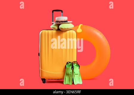 Yellow travel suitcase with summer vacation accessories, inflatable ring, flippers, hat, and sunglasses on red background Stock Photo