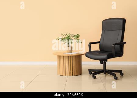 Modern office interior with black ergonomic chair, wooden side table, and potted plant against a peach wall Stock Photo